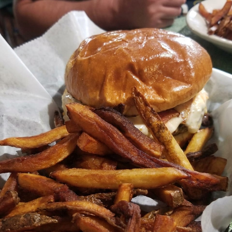 Brioche cheeseburger served with a generous pile of golden fries on parchment paper, with additional fries and a diner’s arm in the background.