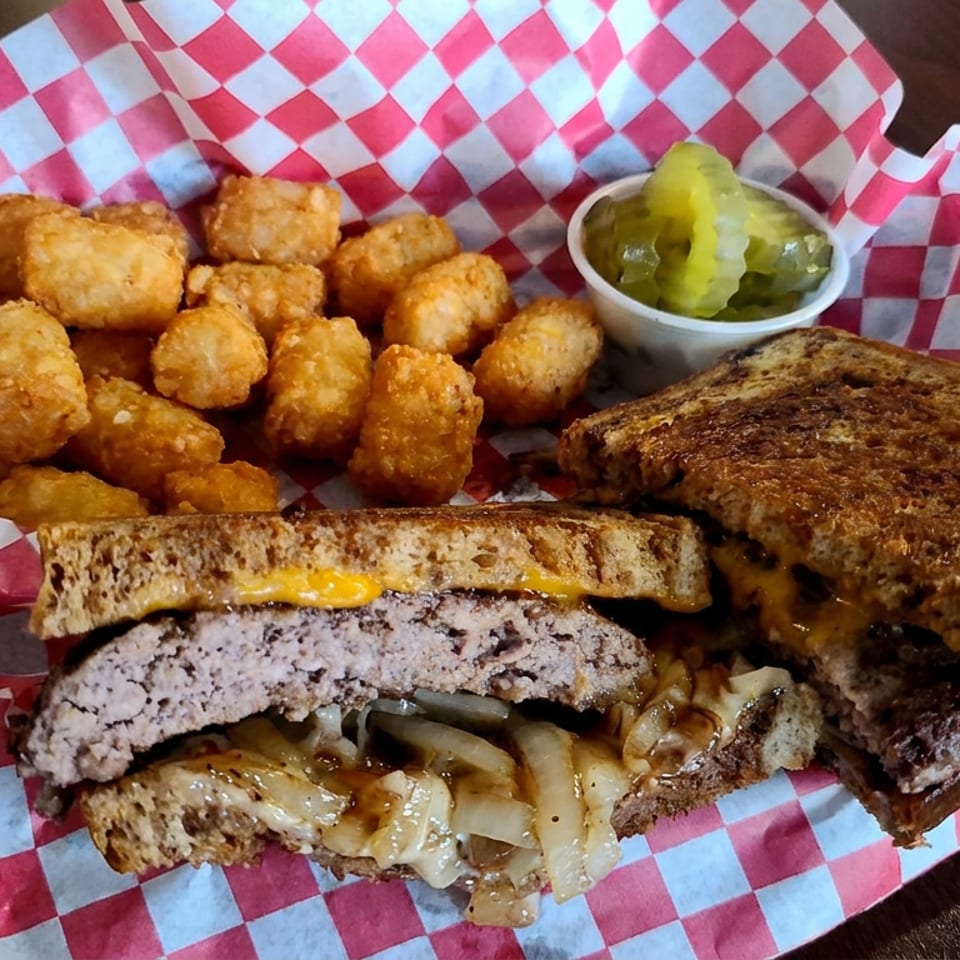 Grilled patty melt with melted cheese and onions, served with golden fried bites and pickles on checkered paper.