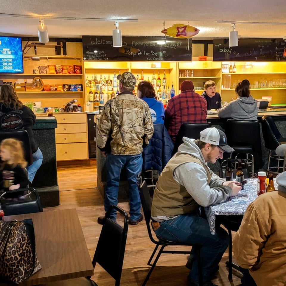 Busy casual restaurant and bar interior with patrons seated at tables, drinks, televisions, and illuminated liquor shelves.