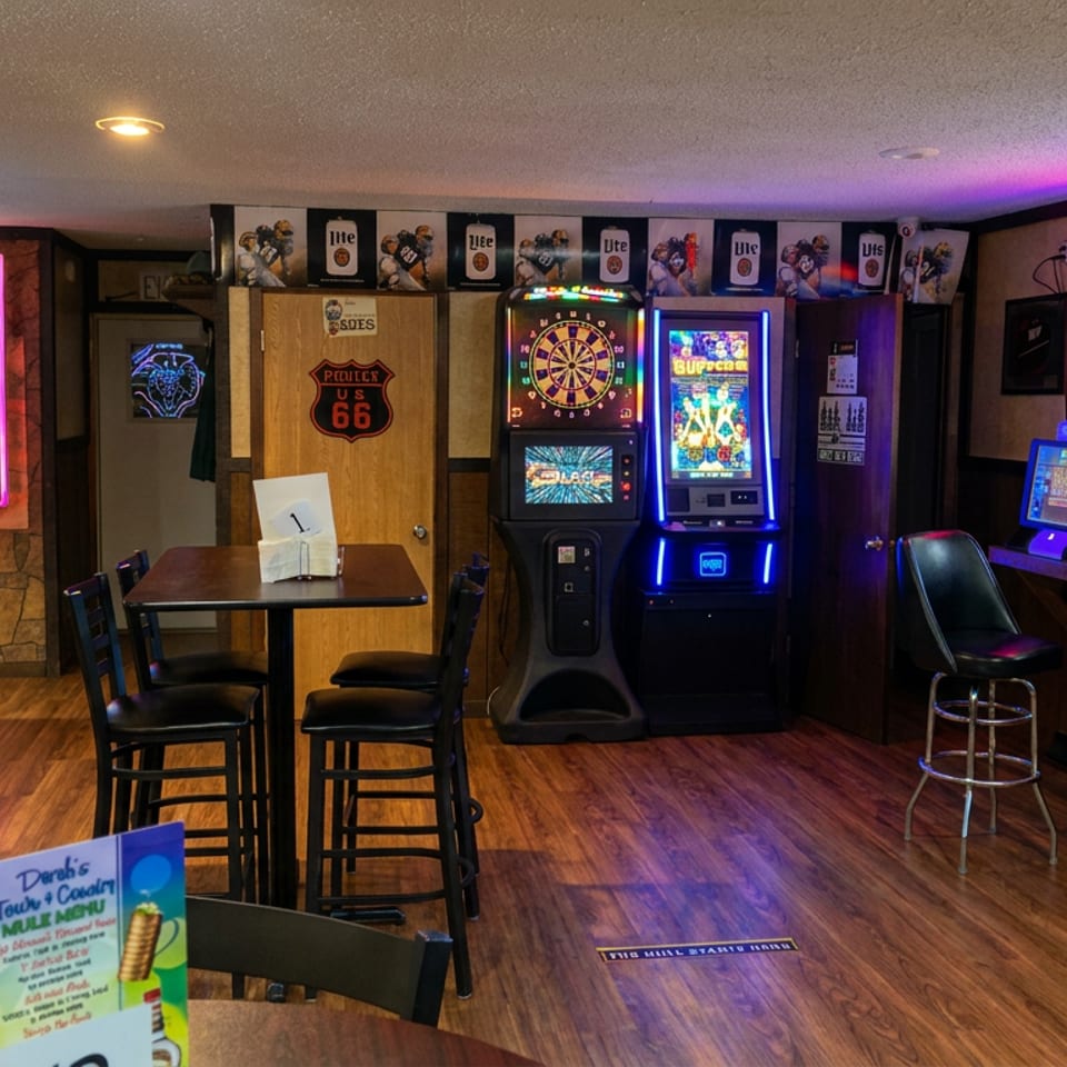 Dimly lit bar arcade with pool table, slot machines, dartboard, high-top tables, and colorful neon beer signs.
