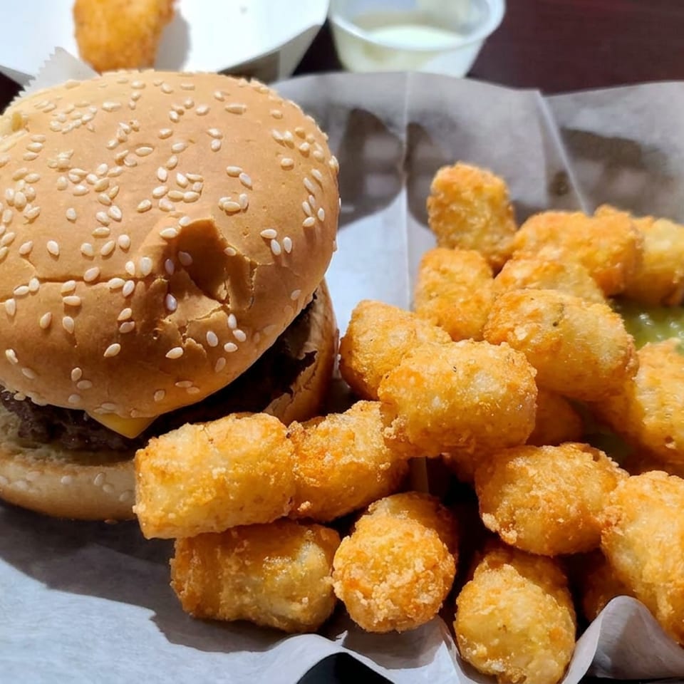 Cheeseburger with a sesame-seed bun and a generous serving of golden fried potato tots, pickles, and dipping sauce on a black tray.