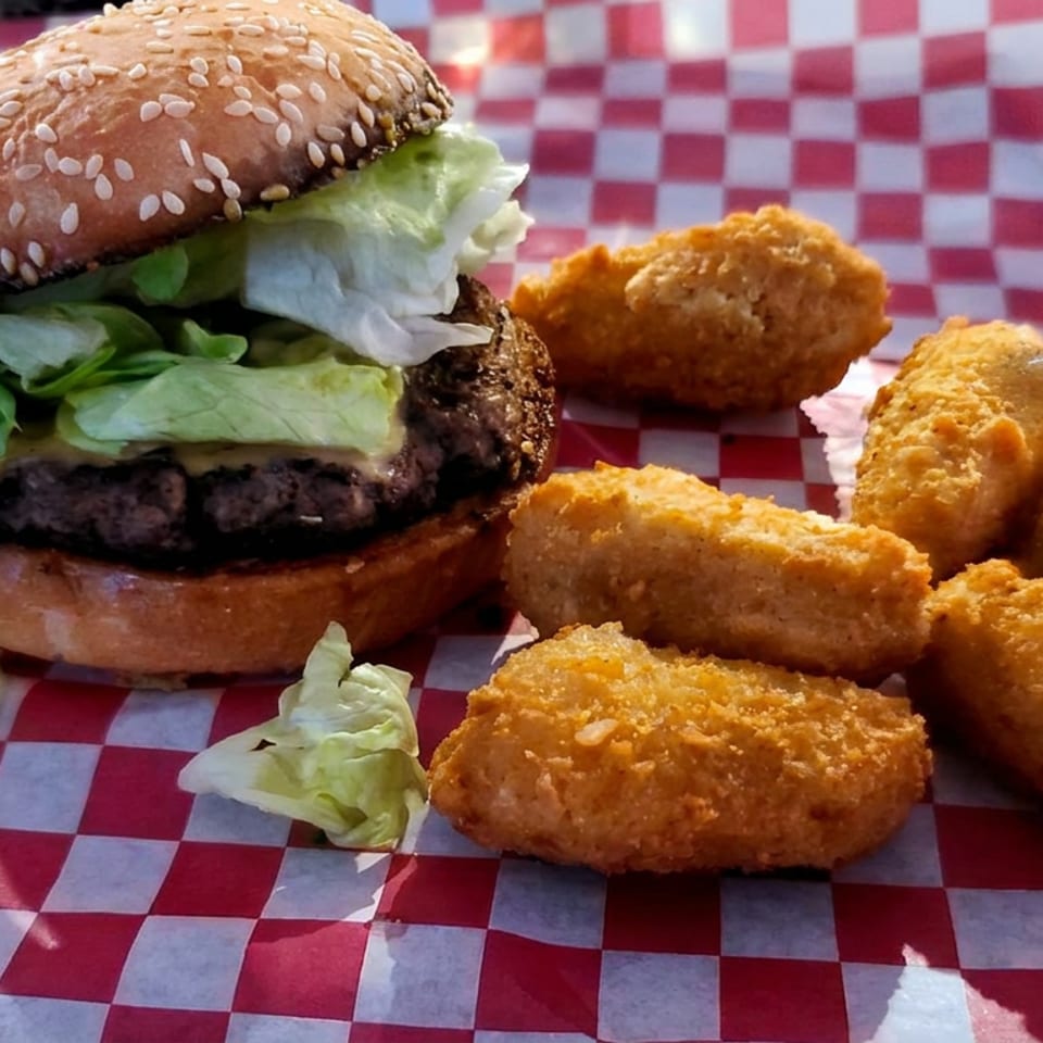 Cheeseburger with lettuce on a sesame bun served alongside several golden fried bites on red-and-white checkered paper.